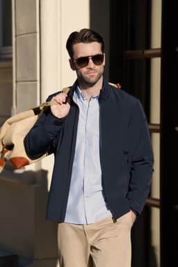 A navy jacket is worn by a man with sunglasses, holding a backpack, standing near a building.