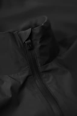 A black jacket featuring a high collar and zip closure, with a minimalist design. It has a logo on the fabric.