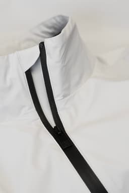 A white jacket with a high collar and a black zipper. Features a minimalistic design and has a logo.