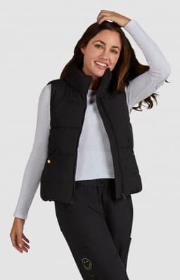 A woman models a black padded vest over a long-sleeve white top, smiling and posing against a plain background.