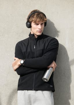 A black men's jacket is worn by a model holding a drink bottle, with headphones on and posing against a grey wall.