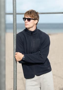 A man wearing a navy polar fleece jacket and sunglasses stands by a beach, leaning against a metal pole.