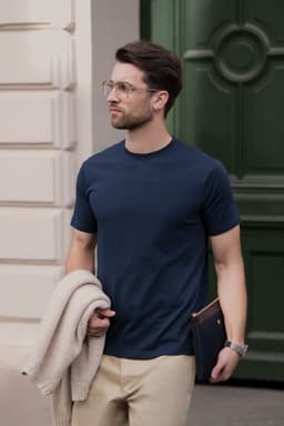 A navy men’s t-shirt is worn by a man holding a beige sweater and a clutch, set against a green door backdrop.