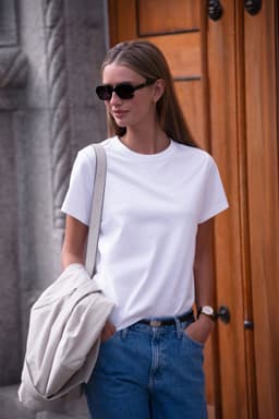 A white women's t-shirt is worn by a model with sunglasses against a wooden background, carrying a light-coloured bag.