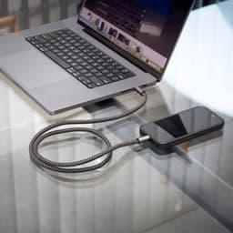 A grey and black charging cable connected to a smartphone and a laptop on a glass table.