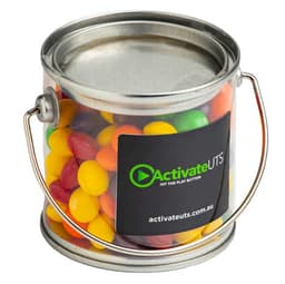 A small clear PVC bucket filled with vibrant Skittles in various colours, featuring a metal handle and a logo.