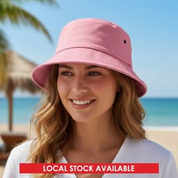 A pink bucket hat sits on a smiling woman's head with a beach backdrop and palm trees. The hat features a logo.