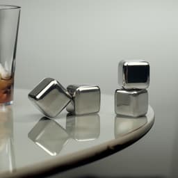 A set of shiny silver whiskey ice cubes resting on a smooth surface, beside a glass of drink.