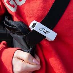 A black pet bag dispenser attached to a strap, featuring a white clip with a logo and a person's hand wearing a red sweatshirt.