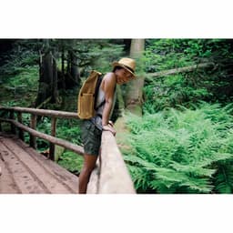 A person wearing a hat and shorts leans on a wooden railing in a lush green forest, with ferns in the background.