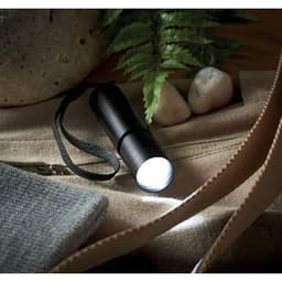 A black flashlight rests on a textured surface alongside pebbles and greenery. It features a strap for easy carrying.