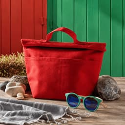 A red tote bag with a handle, placed on a wooden surface alongside sunglasses and stones, with a colorful background.