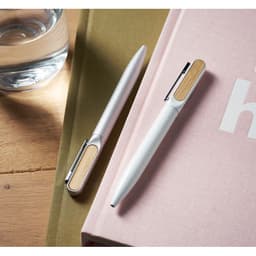 Two pens with bamboo accents, one silver and one white, resting on a pink book beside a glass of water.