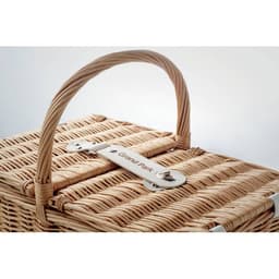 A woven picnic basket in natural tones with a curved handle and a metal clasp. It features a branding label.
