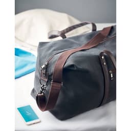 A duffle bag in dark grey with brown zippers and straps, placed on a table beside clothing and a phone.
