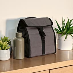 A grey cooler bag with a roll top and metal G hook sits on a wooden surface beside a drink bottle and plants.