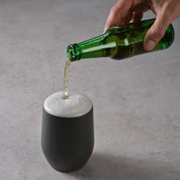 A drink bottle with a matte black finish is being filled with a frothy beverage from a green glass bottle.