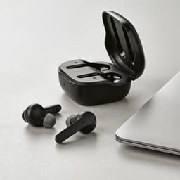 Black wireless earbuds in a charging case, placed next to a laptop on a light-coloured surface.