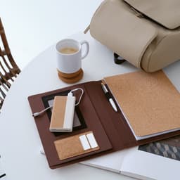 A cork A4 folder with a notepad, power bank, phone, and pen, set on a table with a cup of coffee and a bag.
