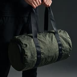 A green gym bag with black straps and a zip closure, held by a person. It features a rounded design and a branding logo.