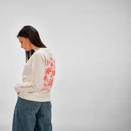 A women's crew neck sweatshirt in cream with a floral design on the back and casual blue jeans. Features a logo.