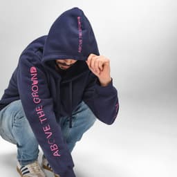 A navy hooded sweatshirt with pink lettering, featuring a logo, worn by a person squatting.