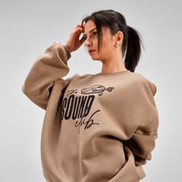 A woman wearing a brown oversized sweatshirt with a logo on the front. The sweatshirt has a relaxed fit and ribbed cuffs.