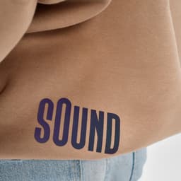A tan oversized sweatshirt made from organic cotton, featuring a large purple text logo. It has a subtle branding detail.