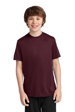 A boy in a burgundy youth performance tee smiles, wearing black shorts. The tee features a logo.