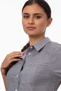 A close-up of a woman wearing a grey classic shirt with a collar and a chest pocket, featuring a logo.