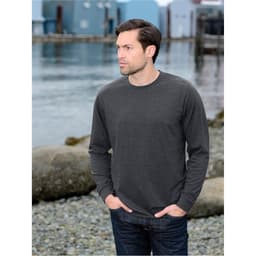 A man wearing a charcoal long-sleeve t-shirt, standing by a waterfront with colourful buildings in the background.