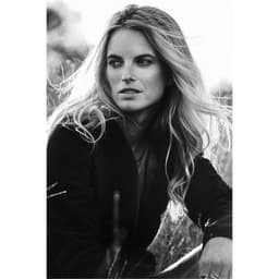 A woman with flowing blonde hair sits amid grass, wearing a black softshell jacket. The image is in black and white.