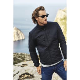 A quilted black jacket with a zip, worn by a man with sunglasses, set against a scenic rocky background.