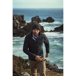 A man wears a navy quilted jacket and a beanie, walking along a rocky shoreline with ocean waves in the background.