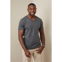 A man in a grey V-neck t-shirt stands against a neutral background, smiling and wearing light-coloured pants.