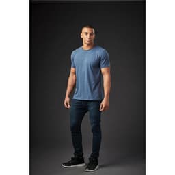 A man models a blue crew neck T-shirt and dark jeans, standing against a dark background. The shirt features a logo.