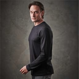A man wearing a black long sleeve t-shirt, posing against a textured background. The shirt features branding.