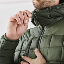 A close-up of a man adjusting the zipper on a dark green puffer jacket. The jacket features a high collar and quilted design.