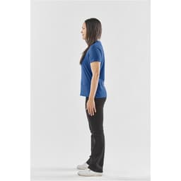 A woman stands in profile wearing a blue T-shirt and black pants, with white sneakers. The image features a logo.