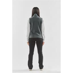A woman stands with her back to the camera, wearing a grey fleece vest over a long-sleeved shirt, paired with black pants.