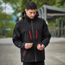 A black softshell jacket with red zippers and a hood, featuring a logo on one side.