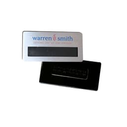 A silver magnetic staff badge with a smooth finish and a black backing. Features a logo.