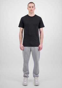 A person wears a black t-shirt and grey track pants, standing against a plain background. The outfit features branding.