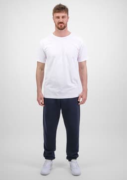 A man stands wearing a white t-shirt and navy blue track pants. The pants have a logo on them.