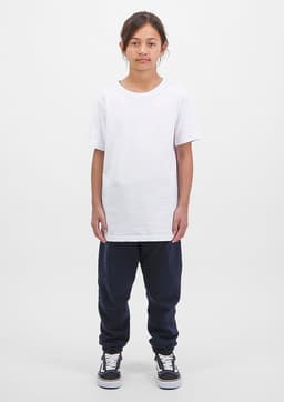 A child wears a white t-shirt and navy track pants, standing against a plain white background. The outfit features a logo.
