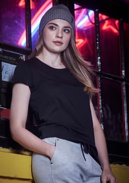 A woman in a black t-shirt and grey pants leans against a window, wearing a grey beanie. The background features neon lights.