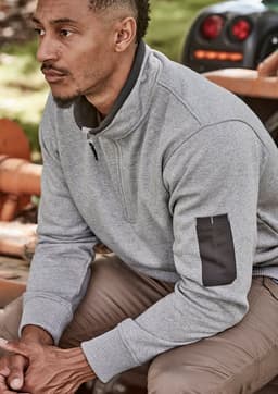 A man wearing a grey unisex 1/4 zip brushed fleece pullover with a logo on the sleeve, seated outdoors.