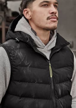 A unisex black hooded puffer vest is worn over a grey hoodie. The vest features a logo and a zip closure.
