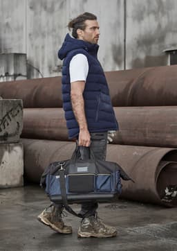A man wearing a navy hooded puffer vest holds a black and navy duffle bag, standing against industrial backdrops.