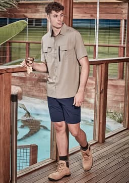 A man wearing a light brown short sleeve button-up shirt and navy shorts, standing on a wooden deck.
